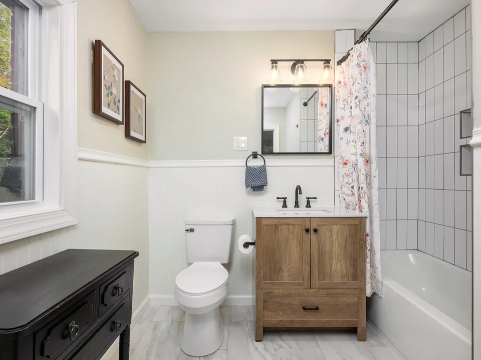 Wood vanity, matte black fixtures, a lighted mirror, beadboard wainscoting and a white-tiled tub and shower.