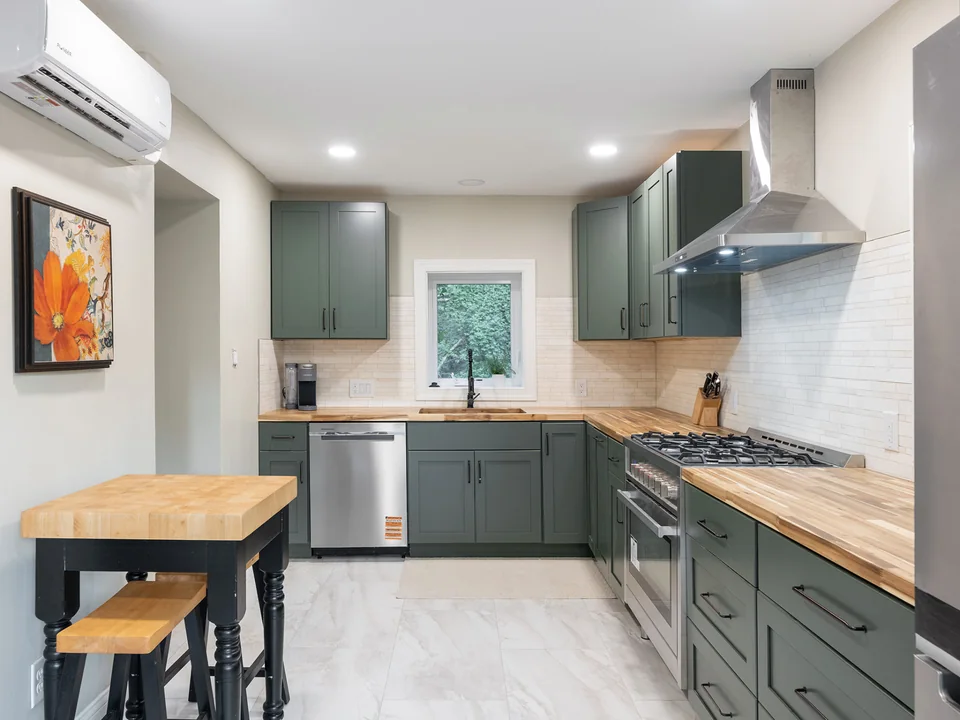 Sage green shaker cabinets, butcher-block counters, stainless appliances, a tile backsplash and a wall-mounted range hood over a stainless gas range.